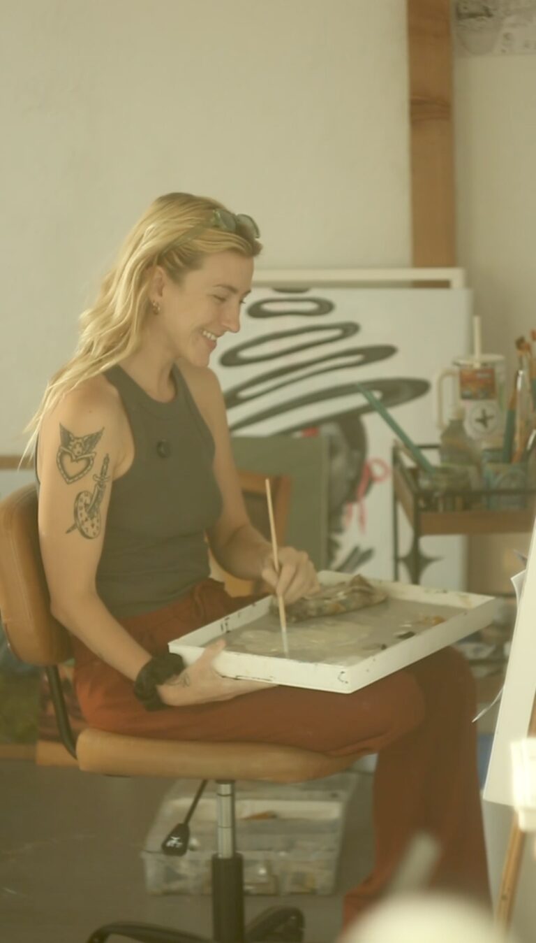 VibeWell Artist-Influencer, Cheyenne Proctor seated with paint brush in hand and a palette on her lap. She is looking at a canvas and smiling, with glasses in her shoulder length blonde hair. She is wearing a dark green tank top and dark orange pants and has two tattoos on her upper arm.
