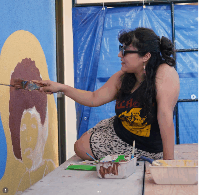 Muralist Elisa Torres sits on a work table outdoors, leaning forward to paint a lmural on a textured wall. She is working on a figure rendered in mauve and white set against a golden oval, with a blue sky visible above. Paint containers, brushes, and a paint tray sit on the table beside her. She is wearing sunglasses, a sleeveless band tee, and a patterned skirt.