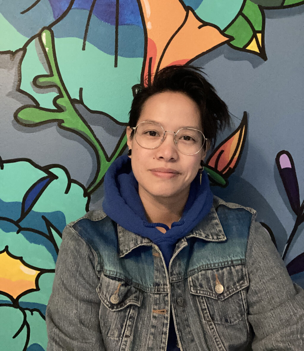 Artist Vanessa Morrow with short dark hair and wire-rimmed glasses, wearing a blue hoodie and denim jacket, smiling softly in front of a large colorful mural. The mural features bold outlined shapes including leaves, flowers, and abstract forms in bright blues, greens, oranges, and purples
